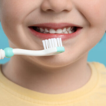 Cute little boy brushing his teeth with plastic toothbrush on light blue background, closeup